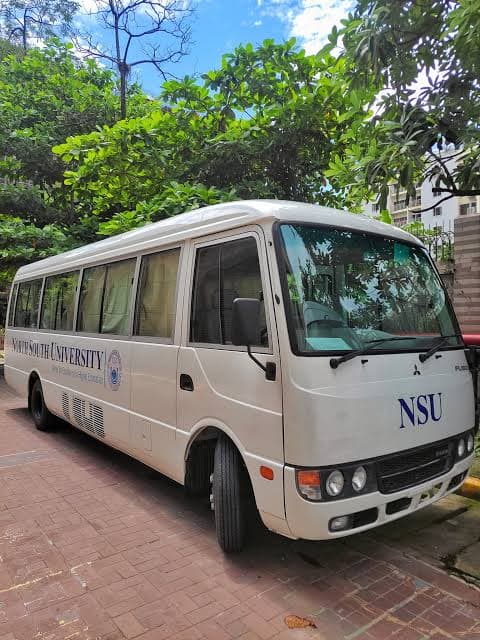 A white North South University minibus with NSU on the front, parked on a brick path under trees